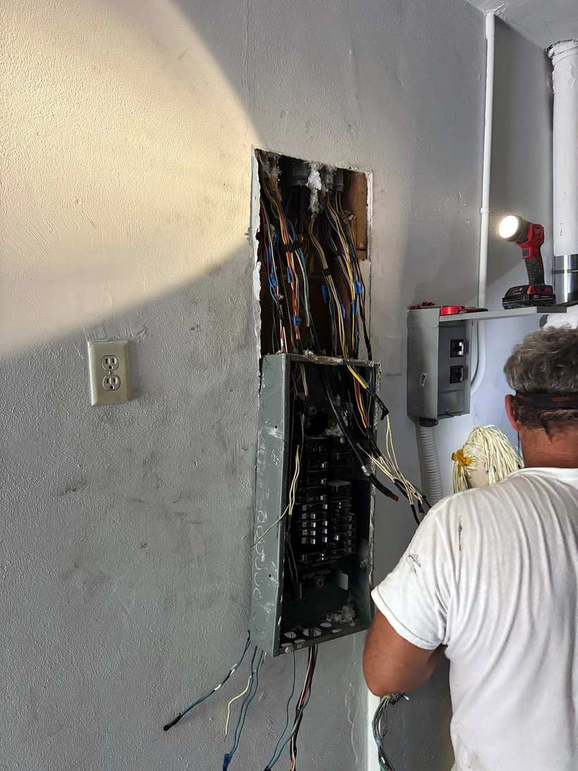 Electrician in a white T-shirt working at an open panel in a wall, wires fanned out around it