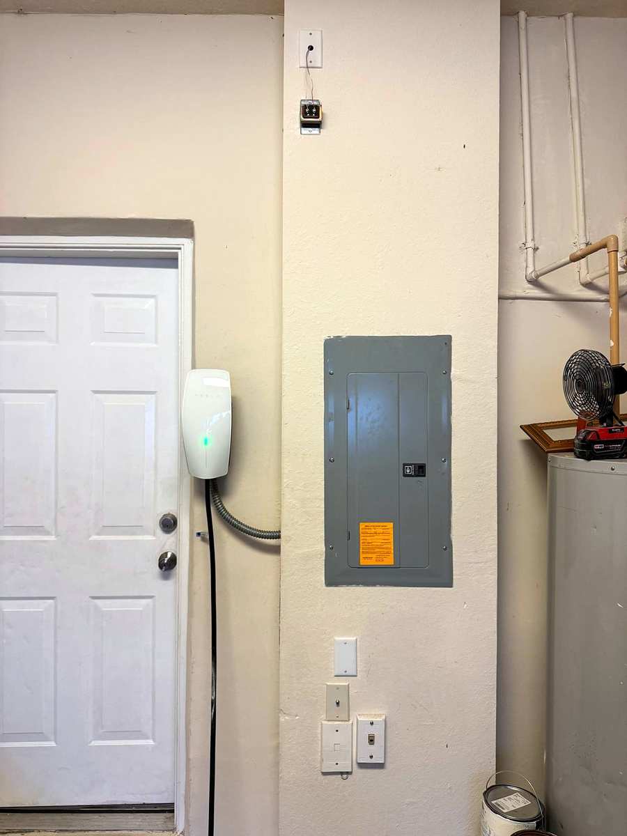 White wall-mounted EV charger with a green status light next to a breaker panel