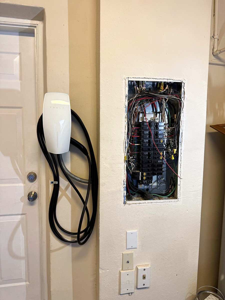 Wall-mounted EV charger with its cable coiled, beside an open breaker panel