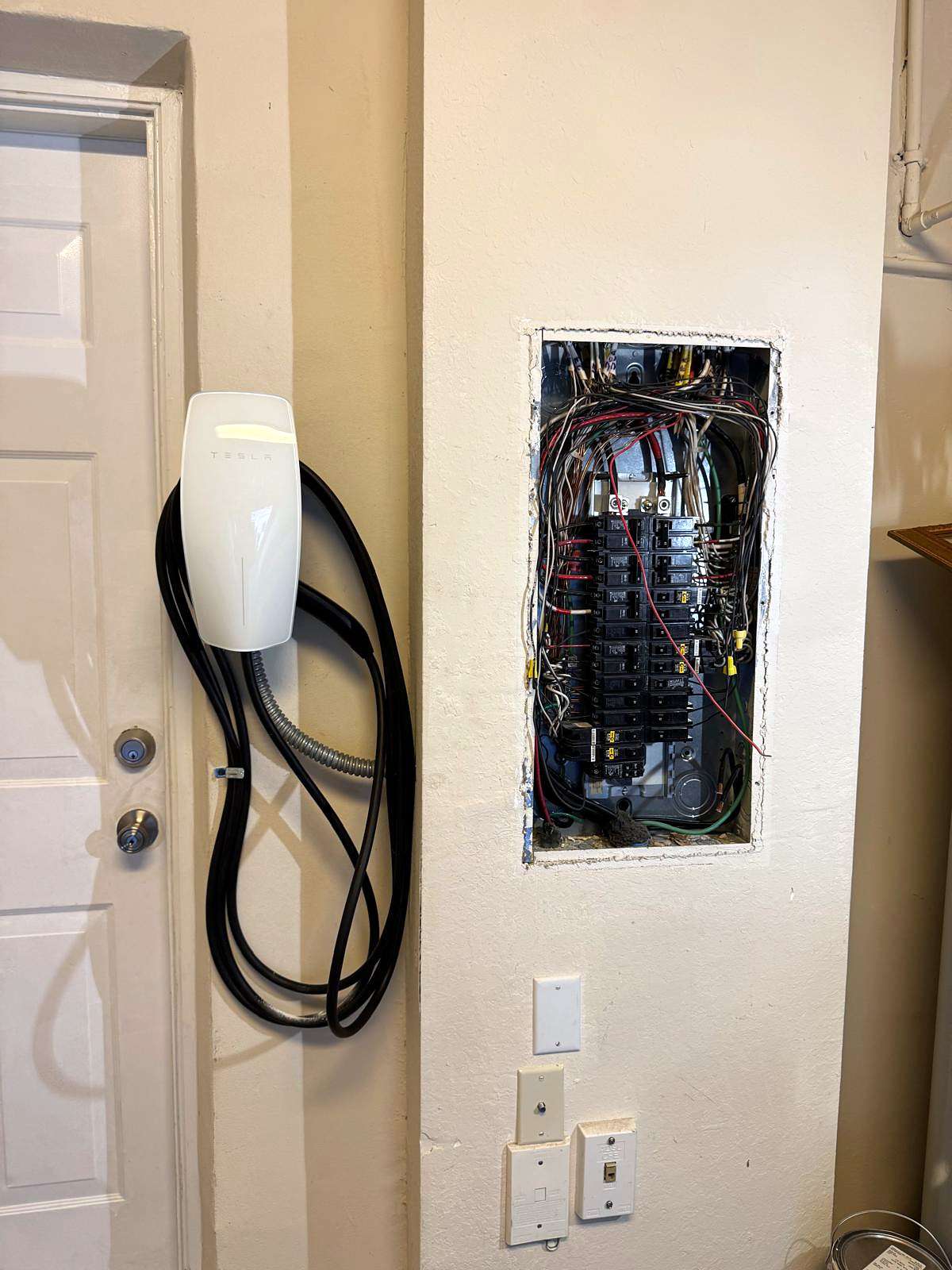 Wall-mounted EV charger with its cable coiled, beside an open breaker panel