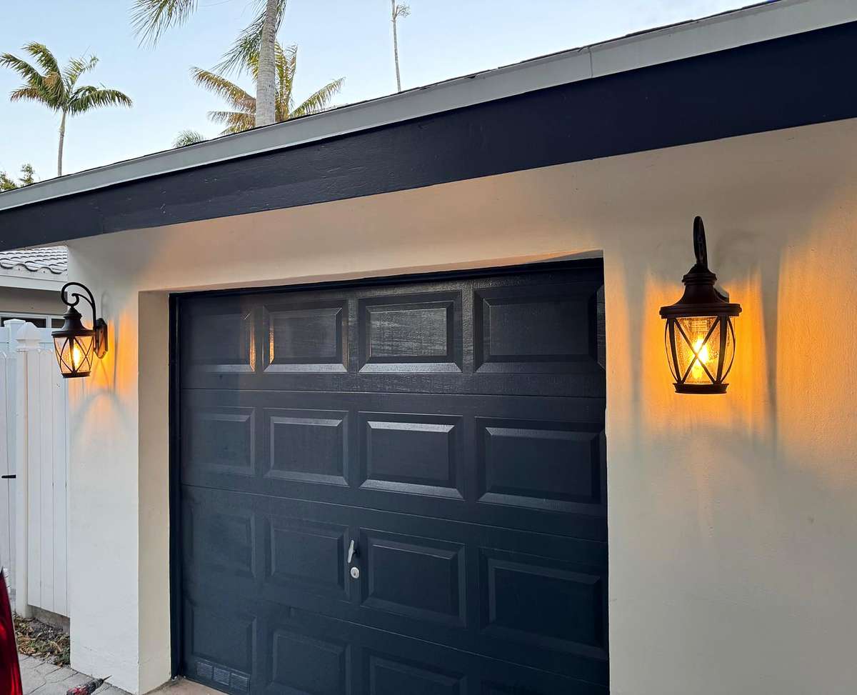 Two lit wall lanterns on either side of a dark garage door at dusk, palm trees above the roofline