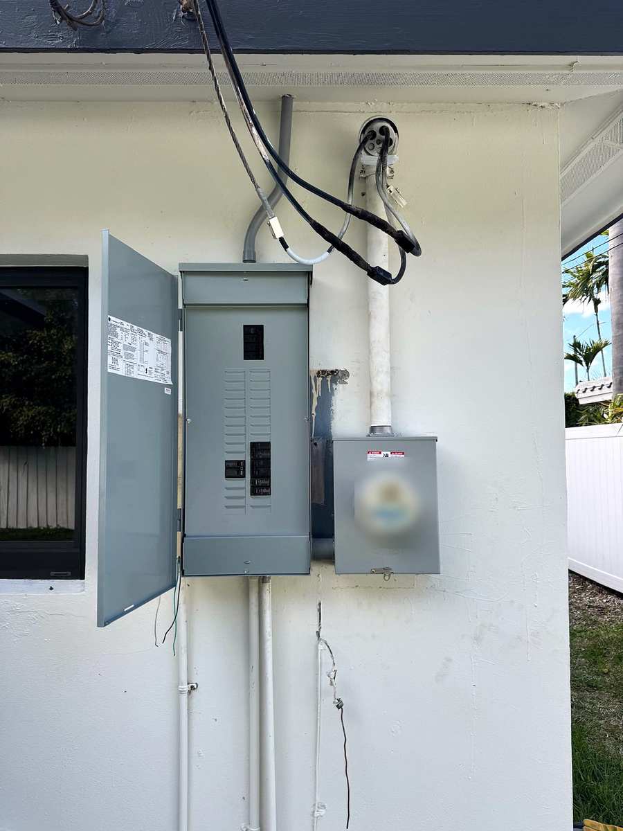 Outdoor electrical service on a white wall: panel with its door open beside the utility meter, palm trees behind