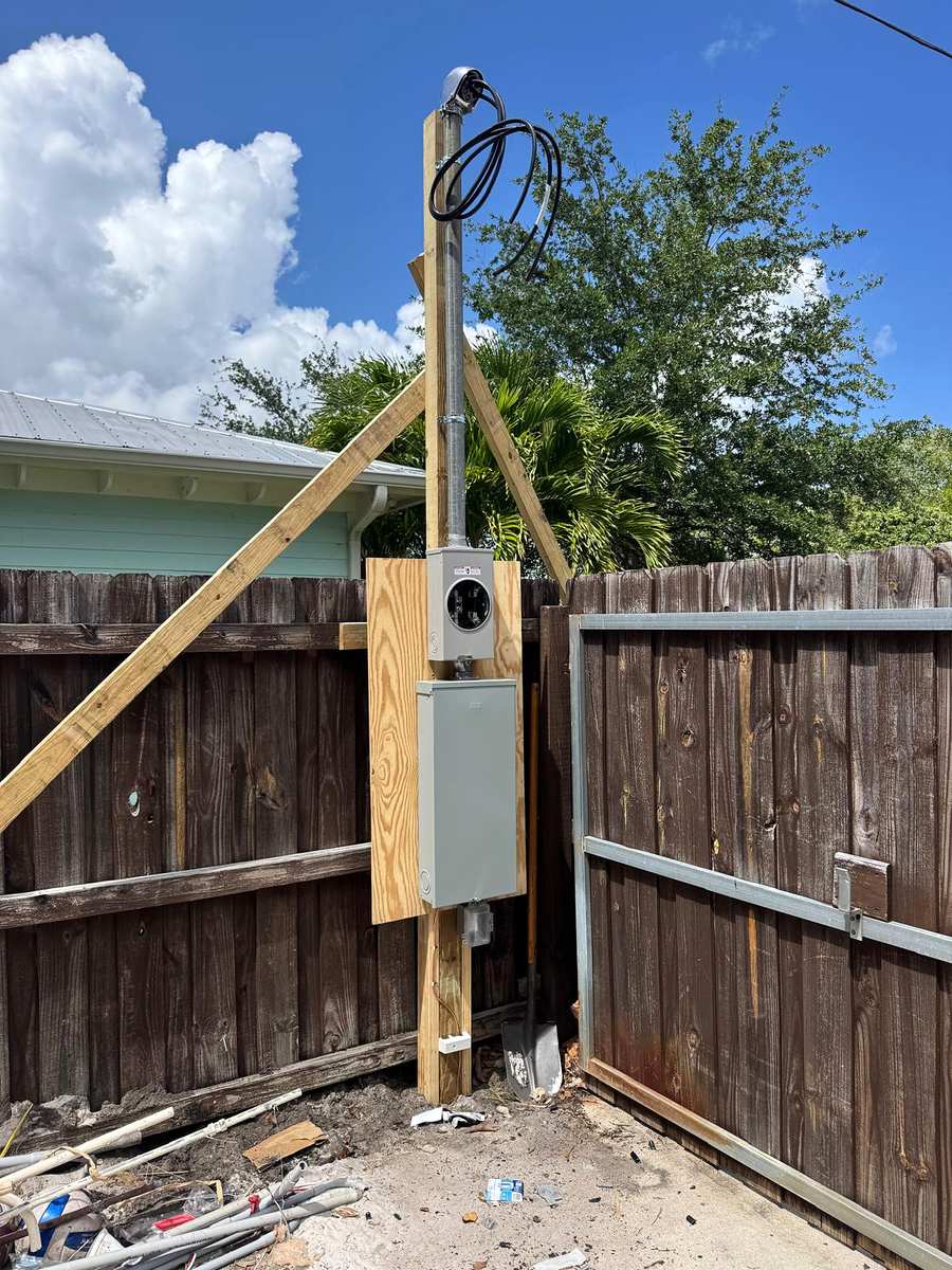Temporary power pole with a meter socket and a small panel, braced with lumber beside a wood fence